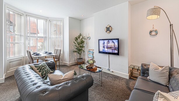 Een moderne woonkamer met een grijze bank, een eethoek met een tafel en stoelen, een aan de muur bevestigde televisie en een groot raam met zonwering.