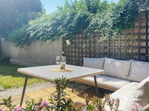 Eine Terrasse mit Tisch, zwei Weingläsern und einer Flasche, umgeben von Grünpflanzen und einem Holzzaun.