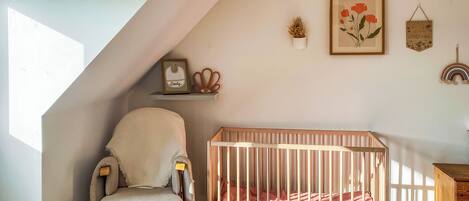 A room with a crib, a cushioned chair, a wooden cabinet, and a sloped ceiling.