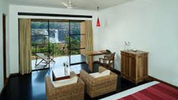 Une chambre d’hôtel avec un grand lit, un bureau, deux fauteuils en osier, une armoire en bois et une vue sur une cascade, visible à travers une porte vitrée.