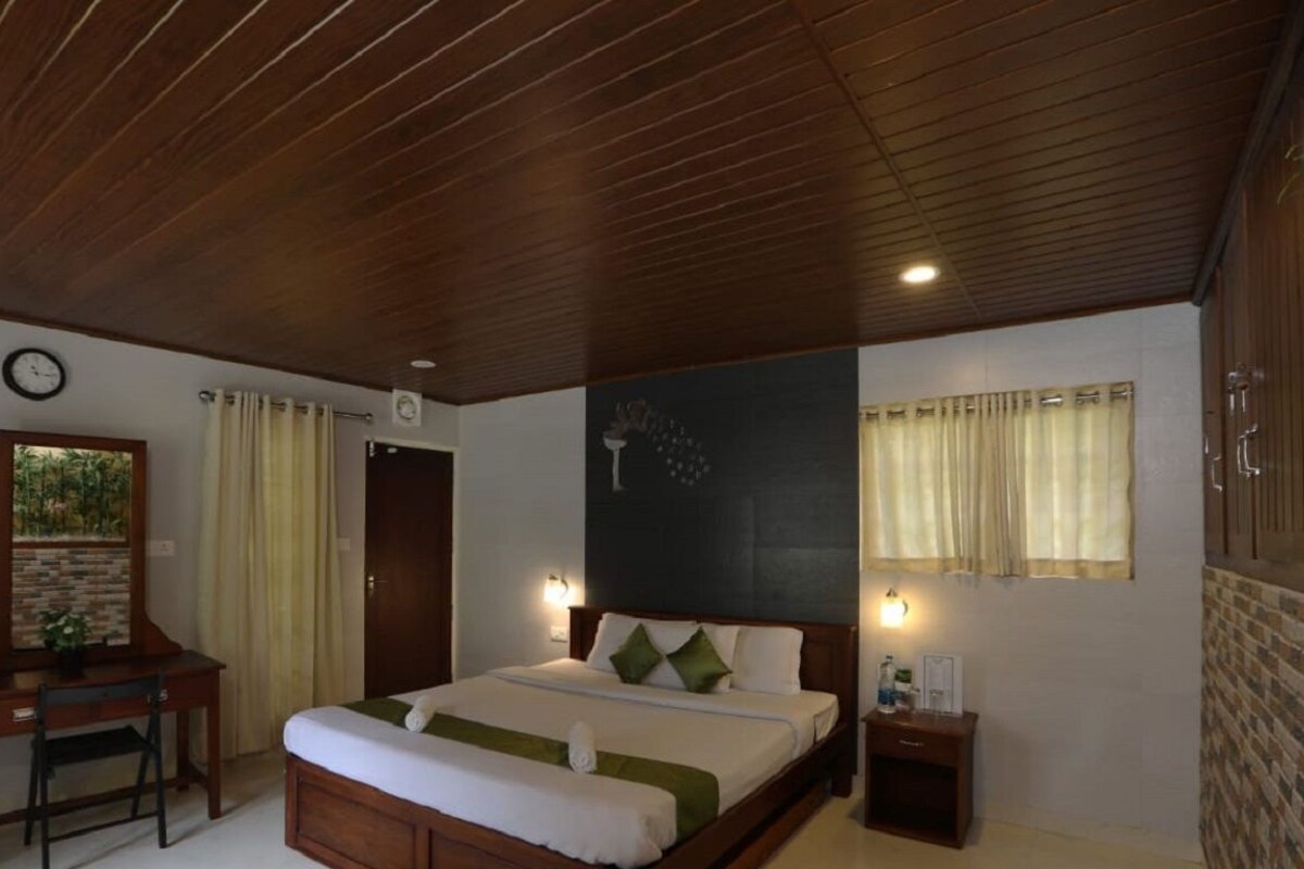 A bedroom with a wooden ceiling, a bed with green pillows, a desk with a chair, a clock on the wall, and a window with curtains.