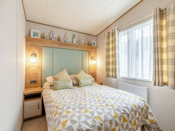 Un dormitorio con cama, mesitas de noche, cabecera decorada y una ventana con cortinas, además de un radiador.