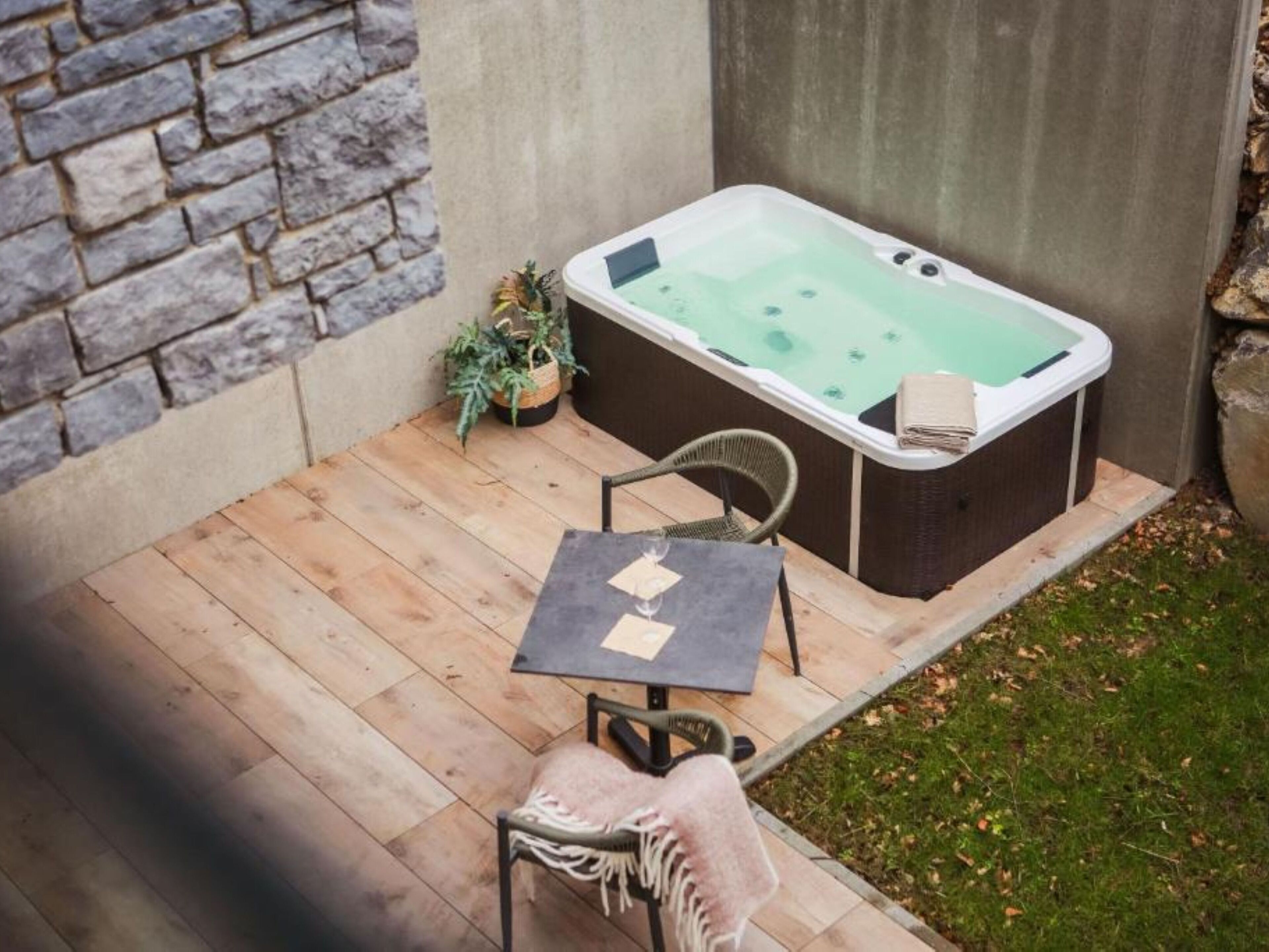 A hot tub in a backyard with a stone wall, a wooden deck, a chair, a table, and a potted plant.