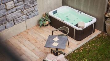 A hot tub in a backyard with a stone wall, a wooden deck, a chair, a table, and a potted plant.