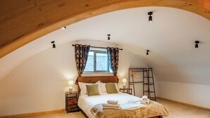 Une chambre avec un plafond en pente, un lit avec des oreillers et une couverture, deux lampes de chevet, une fenêtre avec des rideaux et une échelle en bois appuyée contre le mur.