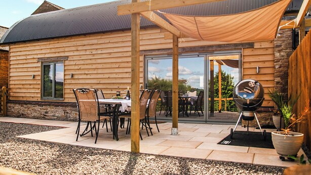 Une cabane en bois dotée d’un patio comprenant une table et des chaises, un barbecue et une pergola offrant de l’ombre.
