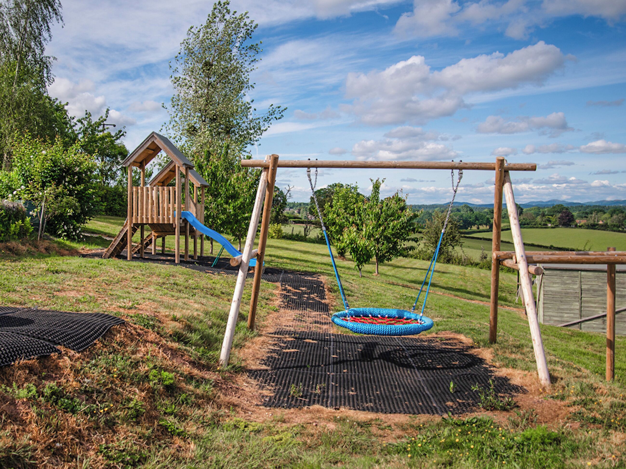 Une aire de jeux avec des balançoires et un toboggan, entourée d’herbe et d’arbres.