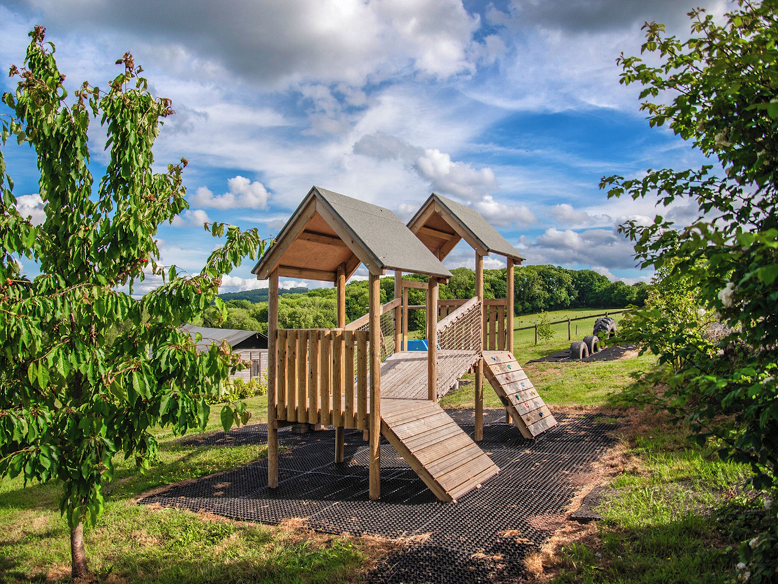A wooden play structure with two slides and a climbing net, set on a grassy area with trees and a clear sky.