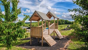 Une structure de jeu en bois, dotée de deux toboggans et d’un filet d’escalade, installée sur une pelouse avec des arbres et sous un ciel dégagé.