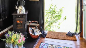A cozy room with a wood-burning stove, a small table with a newspaper, and a window with a view of greenery.