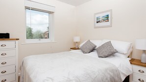 Une chambre à coucher comprenant un lit, une fenêtre, une table de chevet, une lampe, une commode et un tableau encadré accroché au mur.