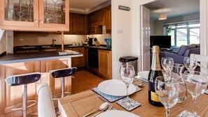 Une cuisine moderne avec des meubles en bois, un coin repas avec une table dressée pour six personnes, et un salon visible à travers une porte ouverte.