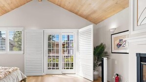 Une chambre avec un plafond en pente, un lit, une table de chevet, une fenêtre avec des stores, une porte avec des panneaux de verre et une cheminée.