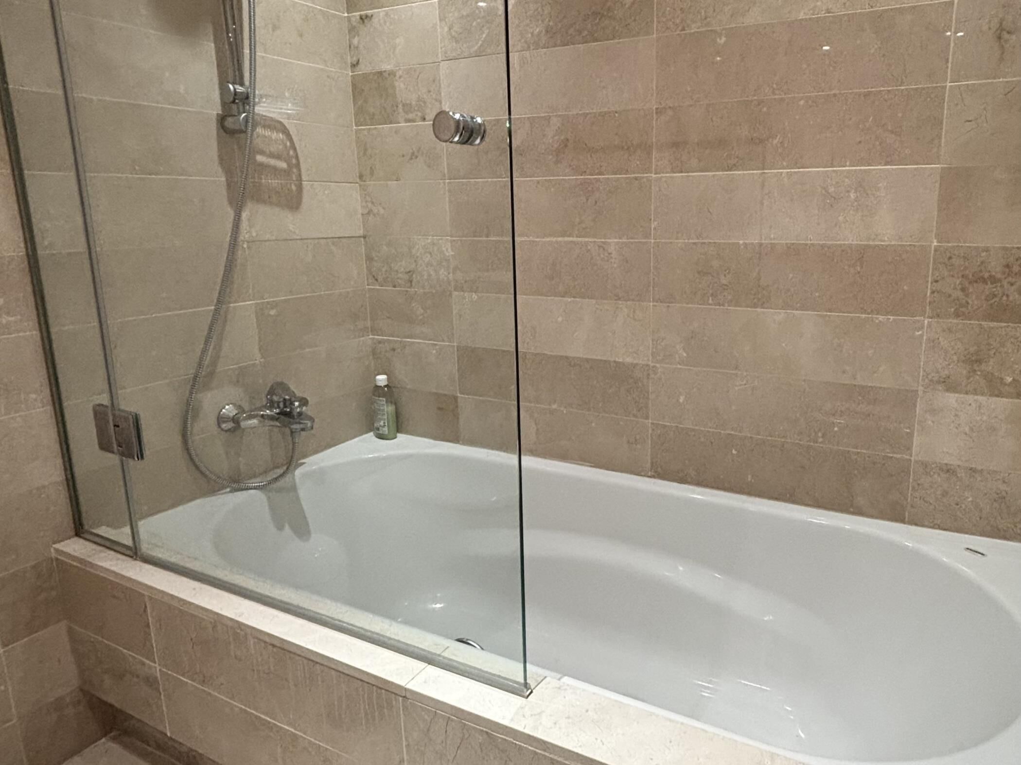 A glass-enclosed bathtub with a showerhead, a handheld shower, and a bottle of shampoo.