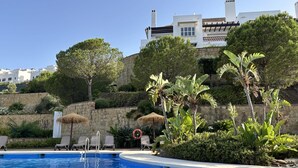 Ein Swimmingpool mit Liegestühlen, eine Steinmauer und im Hintergrund ein Gebäude mit weißen Wänden und Schornsteinen.