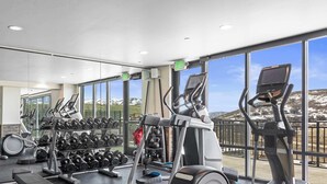 Uma academia com diversos equipamentos de ginástica, amplas janelas com vista para as montanhas e um interior moderno e impecável.
