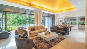 A modern living room with a large window, a sofa, a coffee table, and a dining area. - Luxury4BRVillaPrivatePoolGolf Views v145 (Kathu)