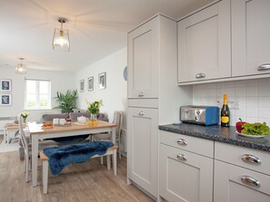 A modern kitchen with white cabinets, a wooden dining table, a blue furry chair, and a countertop with a toaster and a bottle of champagne. - 3 bedroom accommodation in Brixham (Brixham)
