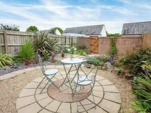 A patio with a round table and chairs, surrounded by a garden with various plants and a wooden fence.