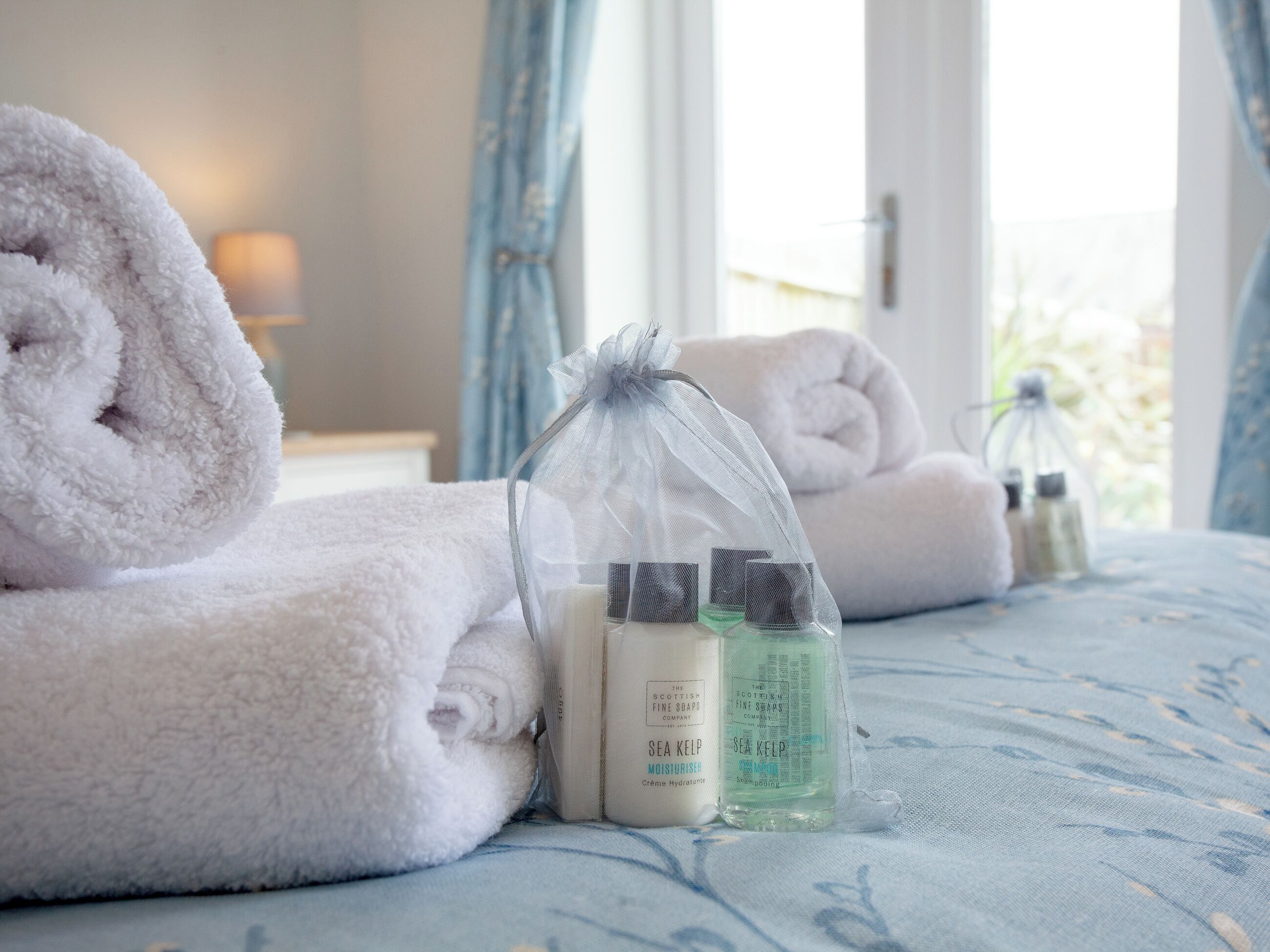 A neatly made bed with rolled white towels, a transparent bag with toiletries, and a window with curtains.