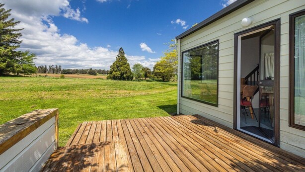 Une terrasse en bois mène à une maison dotée d’une porte-fenêtre coulissante, d’un coin repas avec des chaises et d’une vue sur un champ verdoyant et des arbres.