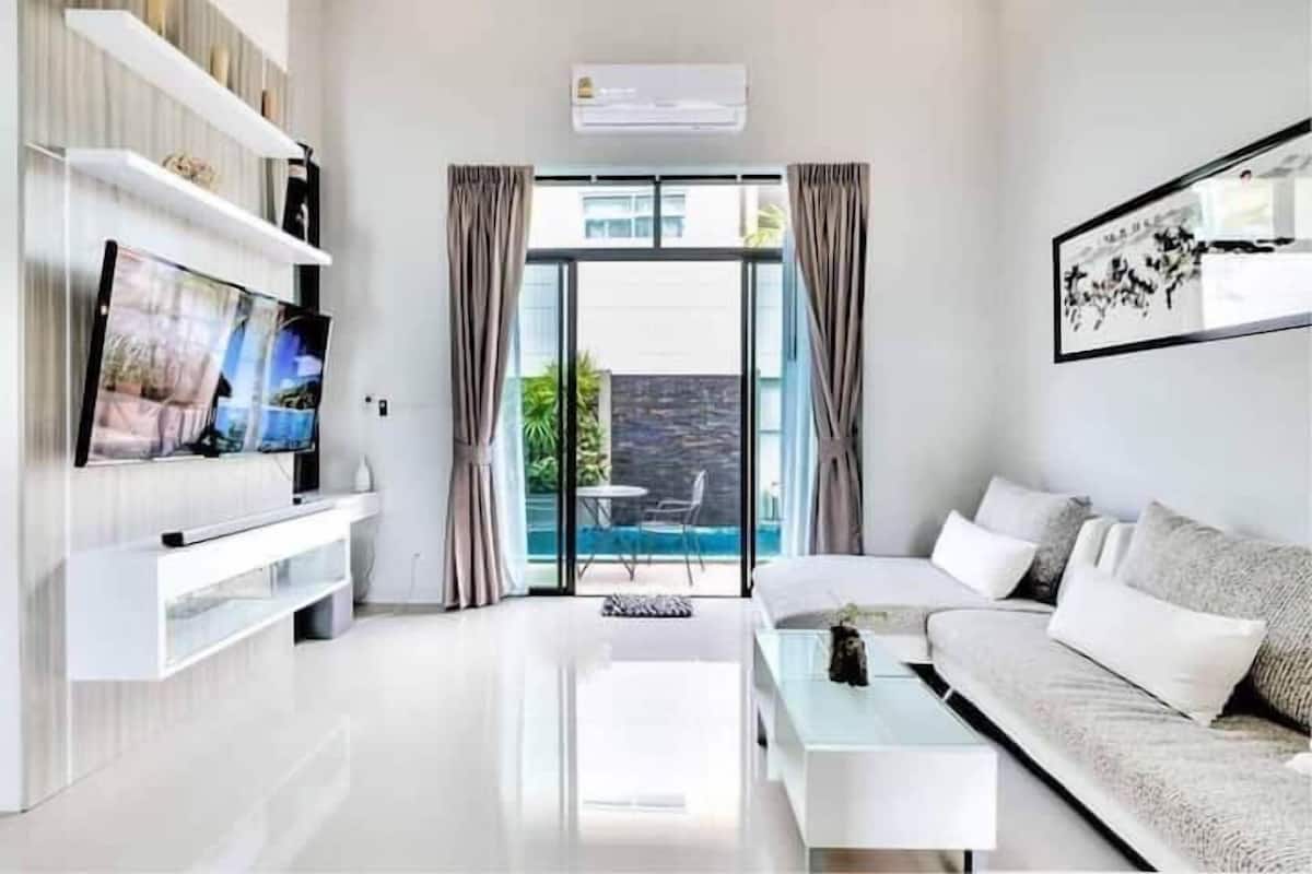 A modern living room with a flat-screen TV, a sofa, a coffee table, and a sliding glass door leading to a balcony with a pool.
