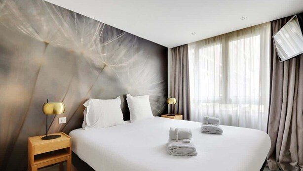 A hotel room with a large bed, two bedside tables with lamps, a window with curtains, and a wall with a textured design. - Appartement cosy - Porte de Versailles (Paris)