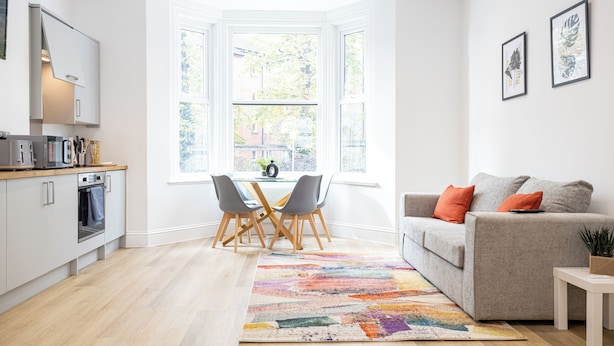 Eine moderne Küche mit weißen Schränken, ein Essbereich mit Tisch und Stühlen, ein graues Sofa mit orangefarbenen Kissen und ein farbenfroher Teppich.