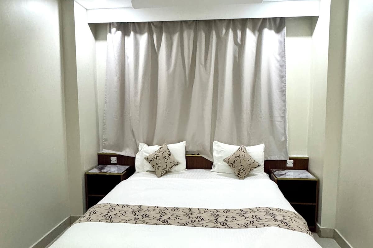 Un lit bien fait, recouvert d’une couvre-lit blanche et agrémenté de deux coussins décoratifs, est placé de part et d’autre de deux tables de chevet sur lesquelles reposent des lampes. La pièce est caractérisée par un plafond et des murs blancs, sans ornements.