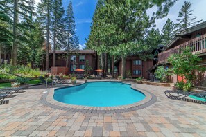 A swimming pool surrounded by a brick patio, with lounge chairs and umbrellas, and a building with a wooden facade in the background. - Walk to Mammoth Village: Chic Mountain Suite 101 (Mammoth Lakes)