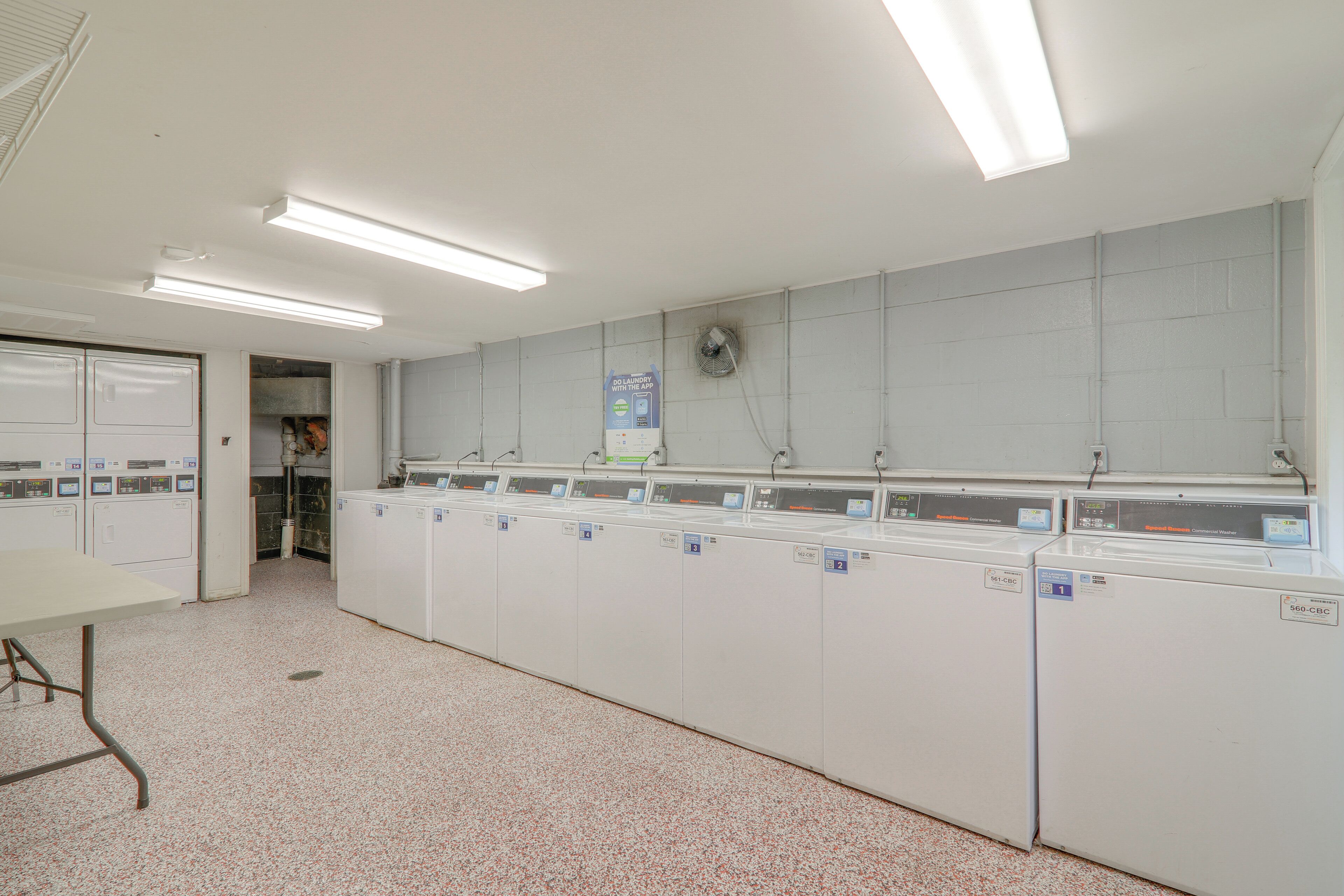 A laundry facility with multiple washing machines and dryers, a ventilation system, and a sign promoting a laundry app.