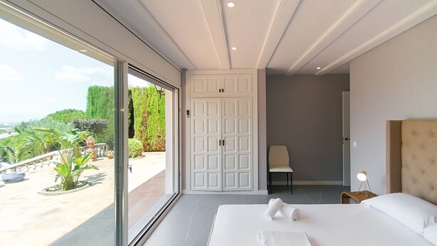 A bedroom with a large bed, a chair, a desk with a lamp, and a sliding door leading to a balcony with plants and a view of the landscape. - WELCS APARTMENT 282 EMP in villa with swimming pool com (Rosas / Roses)