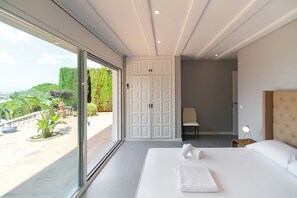 A bedroom with a large bed, a chair, a desk with a lamp, and a sliding door leading to a balcony with plants and a view of the landscape.