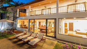 A modern two-story house with a balcony, a pool area with lounge chairs, and a visible interior with a dining area and a bedroom. - CoconutPalmsJacuzzipoolhugeproperty v136 (Rawai)