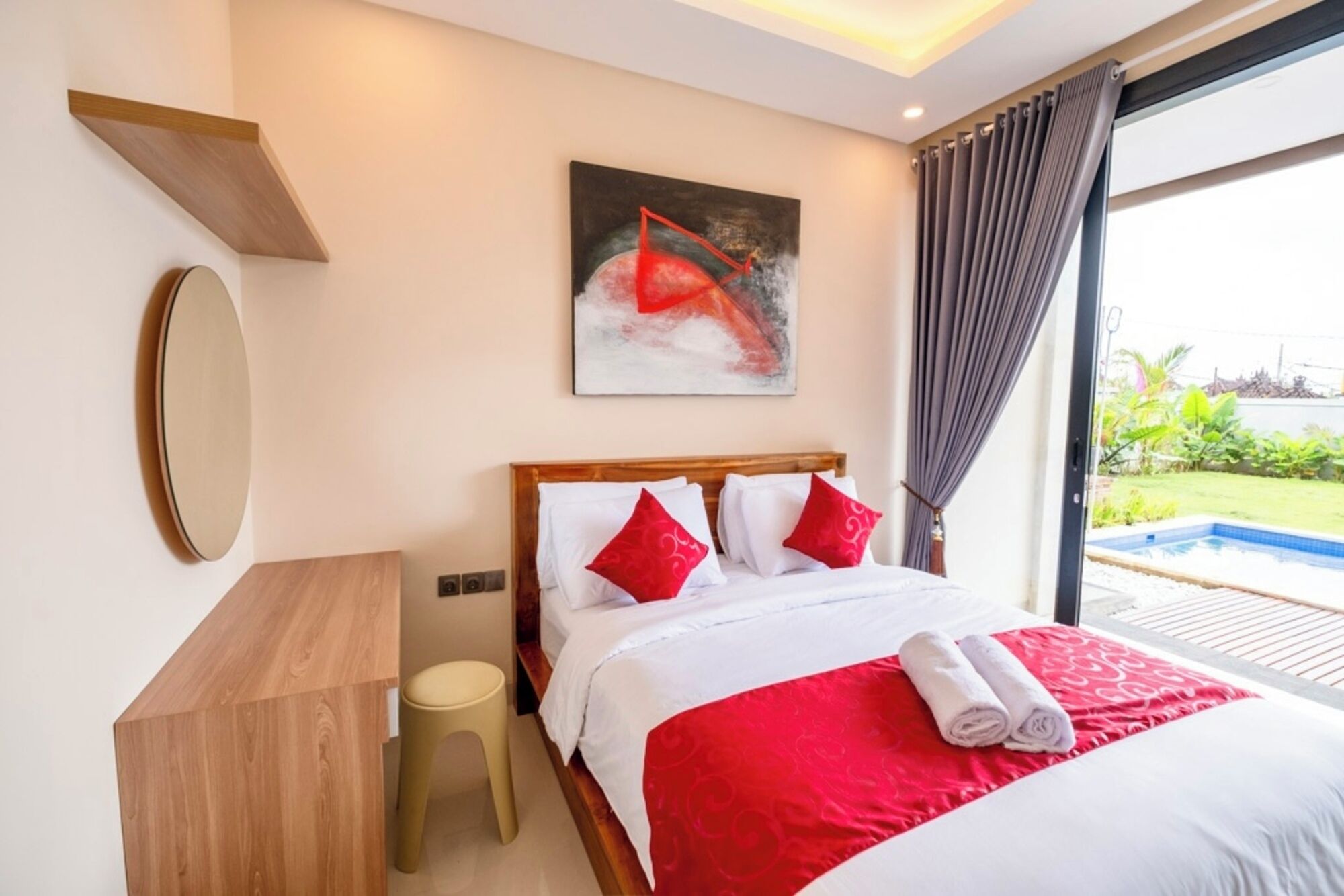 A hotel room with a bed, a wooden nightstand, a stool, a painting, and a view of a swimming pool.