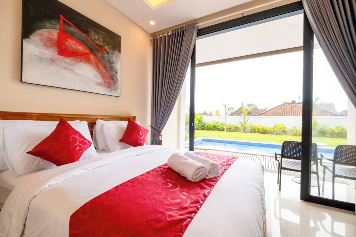 A hotel room with a large bed, red and white bedding, a painting on the wall, and a view of a swimming pool and greenery outside.