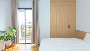 A bedroom with a bed, a wooden wardrobe, a potted plant, and a balcony with curtains. - LUMI WEST LAKE APARTMENT (Hanoi)