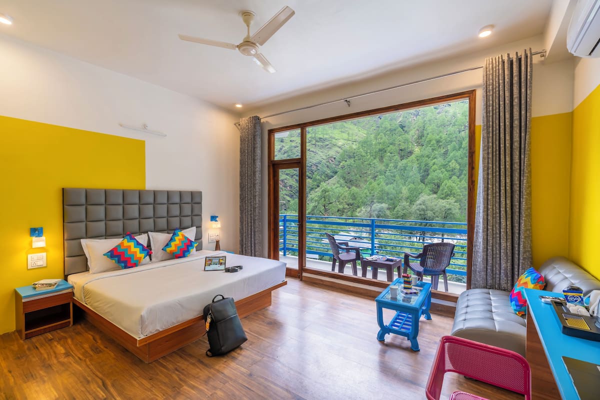 A hotel room with a large bed, a balcony with a view of greenery, a desk with a chair, and a sofa.