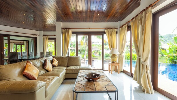A spacious living room with a wooden ceiling, a beige sofa, a coffee table, and large windows offering a view of a swimming pool and greenery. - MassiveHugePool9MinutesWalk to BeachV131 (Choeng Thale)