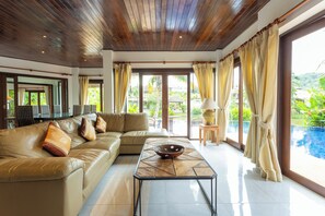 A spacious living room with a wooden ceiling, a beige sofa, a coffee table, and large windows offering a view of a swimming pool and greenery. - MassiveHugePool9MinutesWalk to BeachV131 (Choeng Thale)