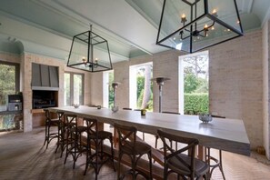 A long dining table with chairs, a modern chandelier, and large windows with a view of greenery. - French Elegance in Bishopscourt (Capetown)