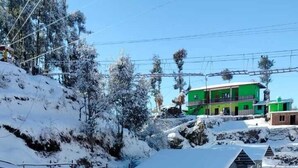 A snow-covered outdoor area with a metal fence, a small building, and a green house in the background. - Snow Adventure Resort (Dhanaulti)