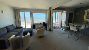 A spacious living room with a large sofa, a dining area with a table and chairs, and a balcony with a view of the ocean. - Apartment in Nieuwpoort-bad With Sea View (Nieuwpoort)