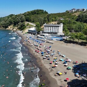 Una playa con numerosos sombrillas y gente, un edificio con piscina, y una colina con árboles.