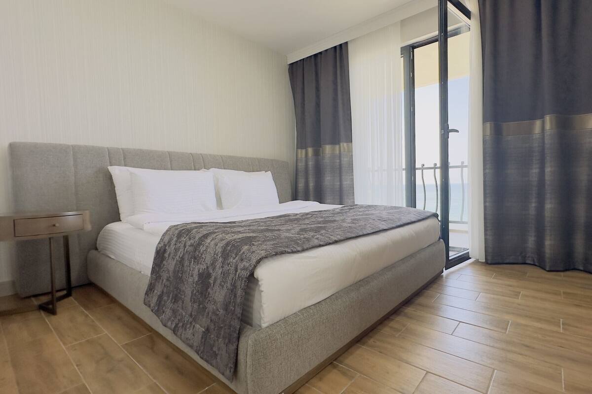 A modern hotel room with a large bed, a bedside table, and a view of the sea through a balcony.