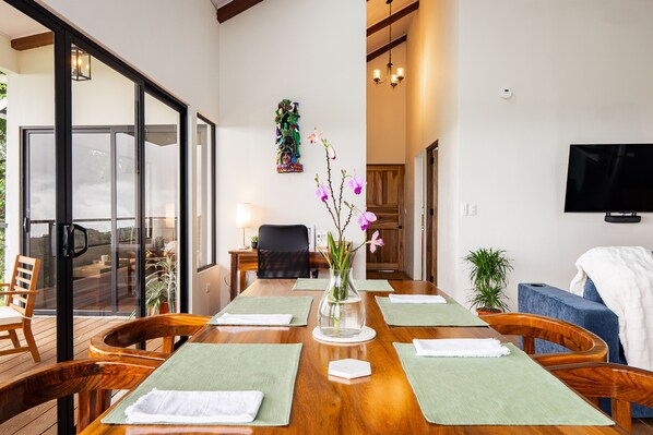 A dining area with a wooden table set for four, a vase with flowers, and a view of the outdoors through large glass doors.