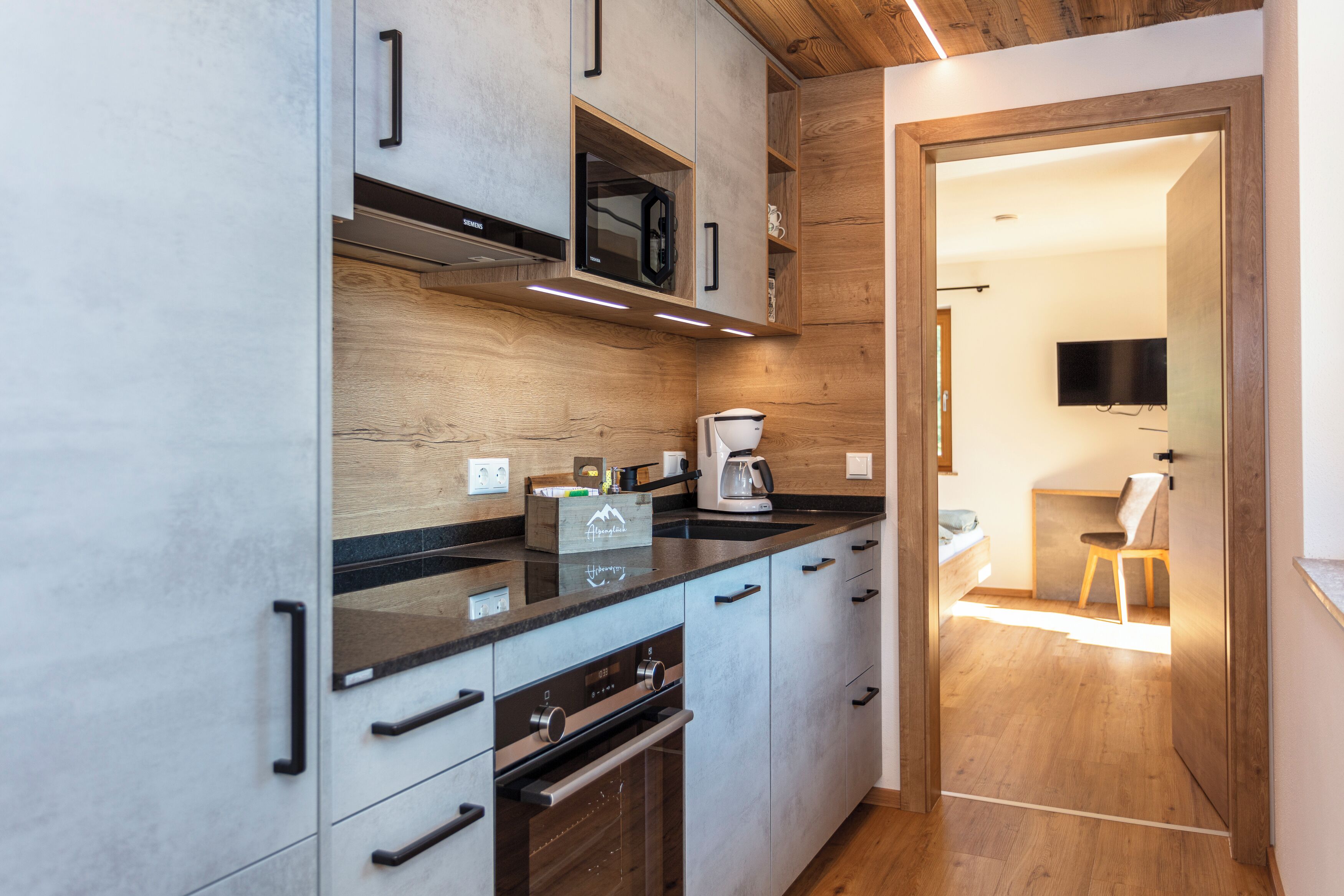 A modern kitchen with built-in appliances, a wooden accent wall, and a dining area visible through an open door.