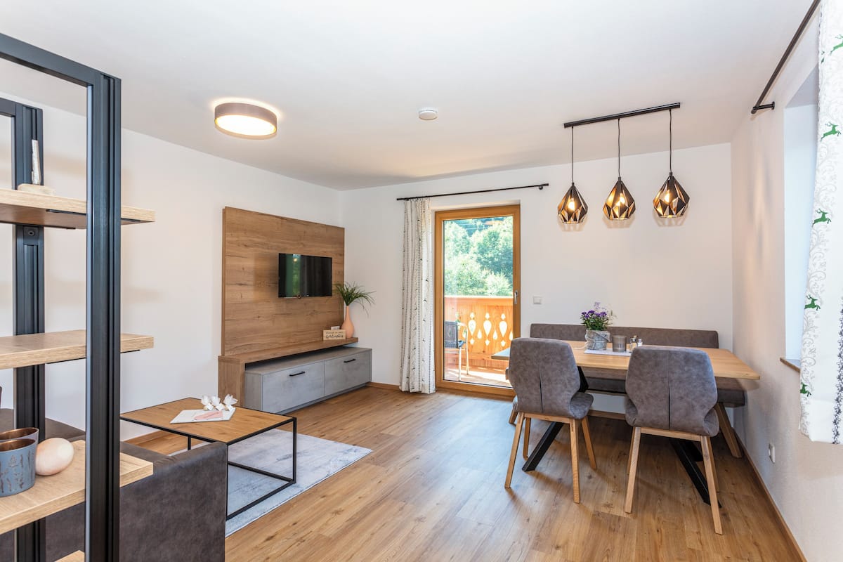 Ein modernes Wohnzimmer mit Essbereich, Holzboden, einem an der Wand befestigten Flachbildfernseher und einer Glastür zum Außenbereich.