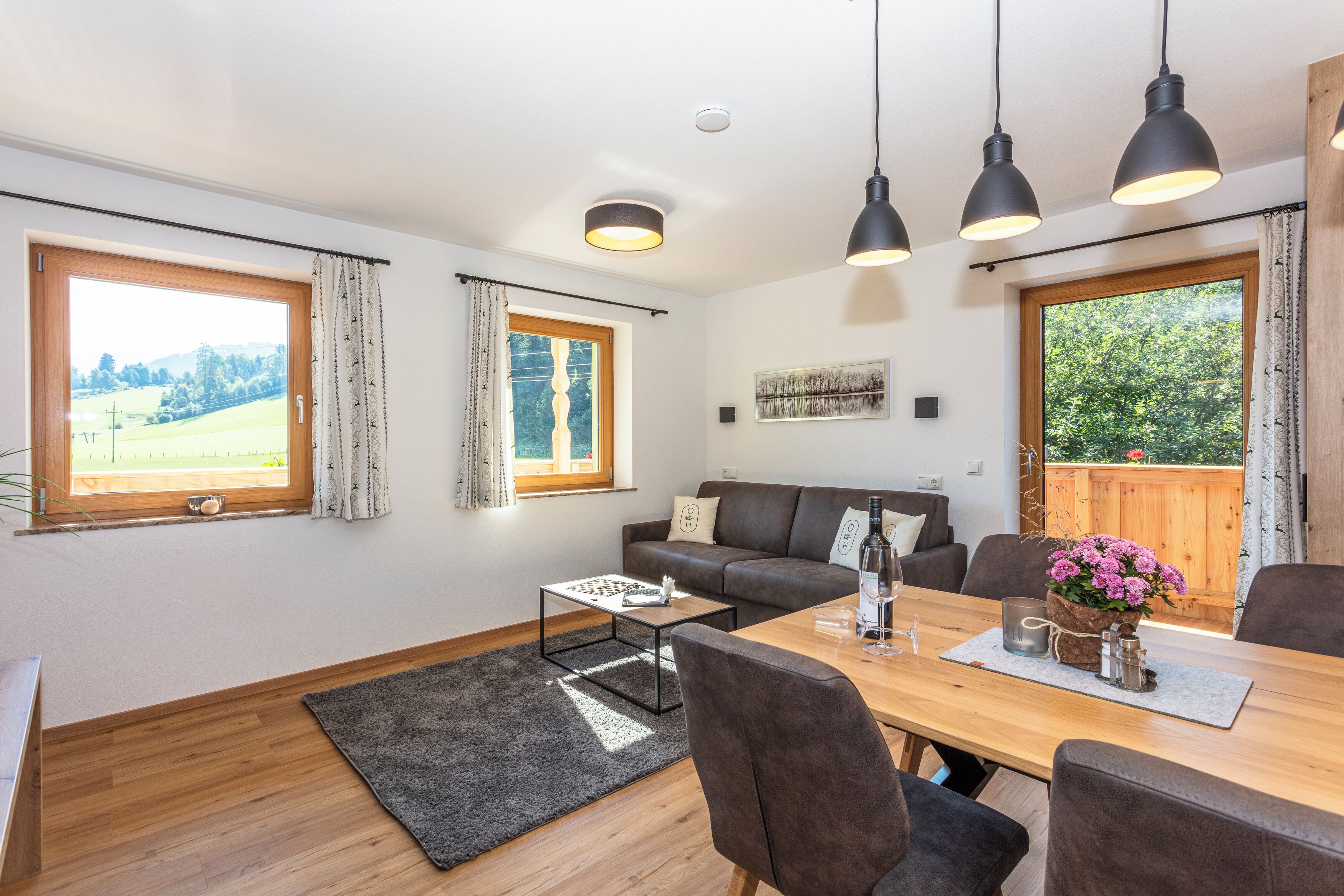 A modern living room with a wooden dining table, a sofa, and a view of a green landscape through large windows.