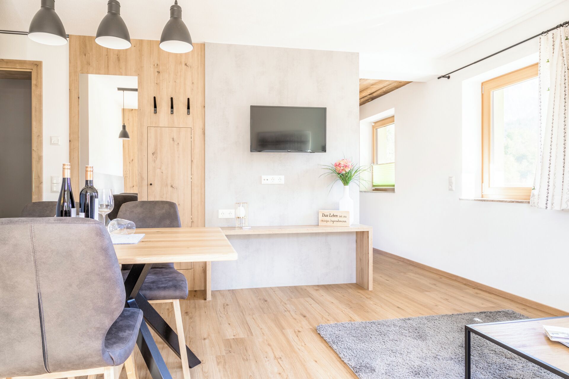A modern kitchen with wooden cabinets, a dining area with a table and chairs, a flat-screen TV mounted on the wall, and a window with curtains.